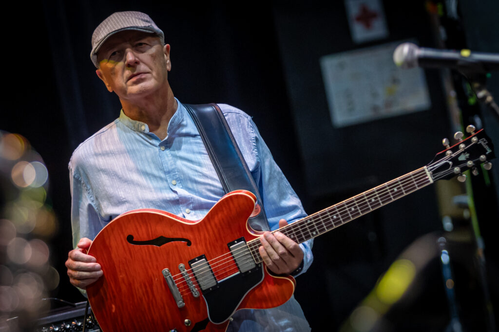 Zdjęcie. Mężczyzna trzyma czerwoną gitarę, którą powiesił na szyi. Mężczyzna ma niebieską koszulę i czapkę z daszkiem, tzw. kaszkiet.