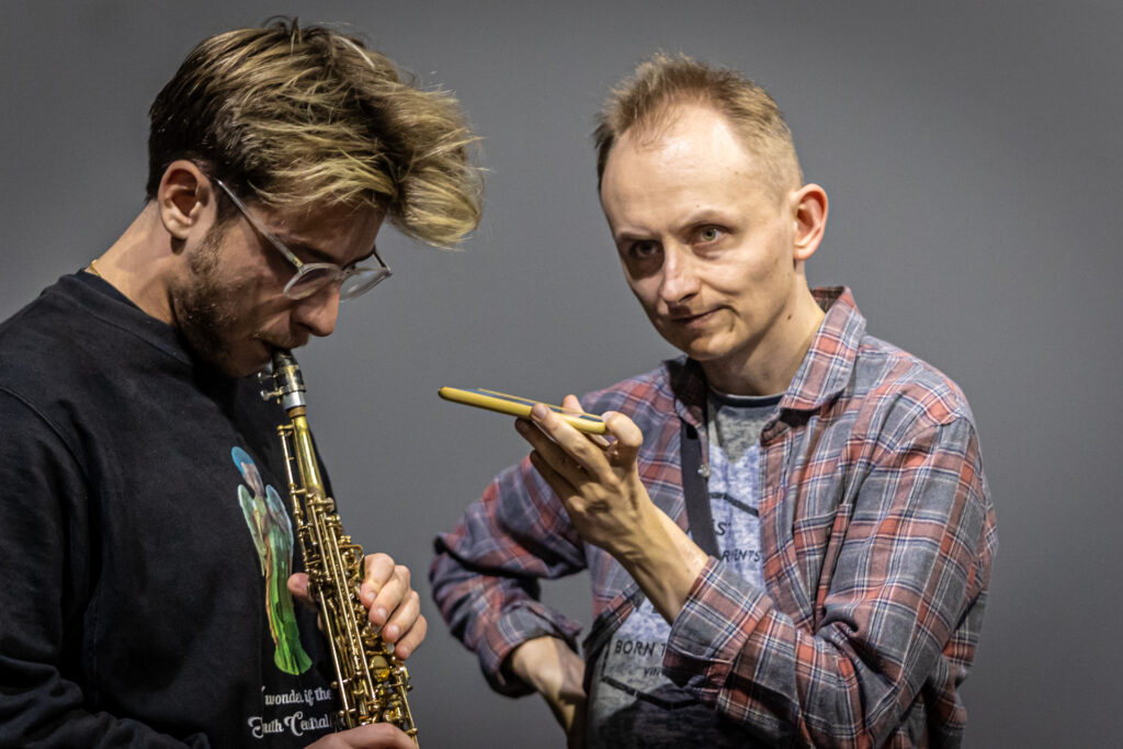 Zdjęcie. Mężczyzna gra na instrumencie dętym. Obok drugi trzyma przy nim telefon komórkowy.