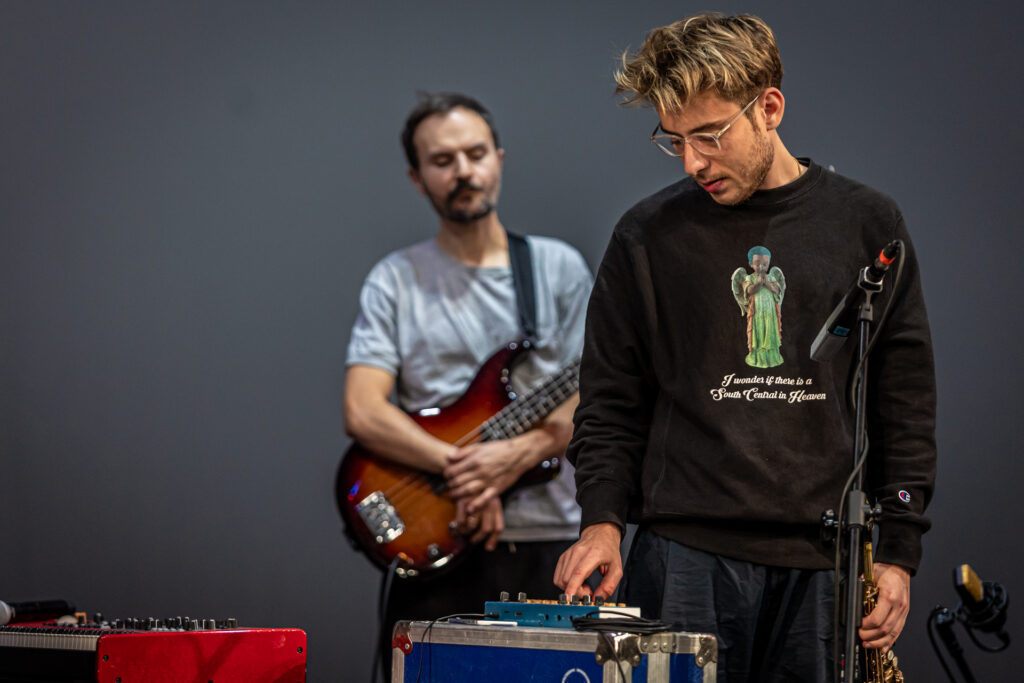 Zdjęćie. Mężczyzna w czarnej bluzie z aniołem w zielonej sukience naciska przyciski miksera. W tle mężczyzna obejmujący swoją gitarę.