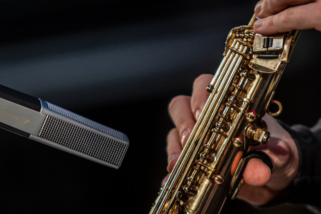 Zdjęcie. Zbliżenie na częśc złotego instrumentu dętego i kwadratowego mikrofonu zbierającego dźwięk.