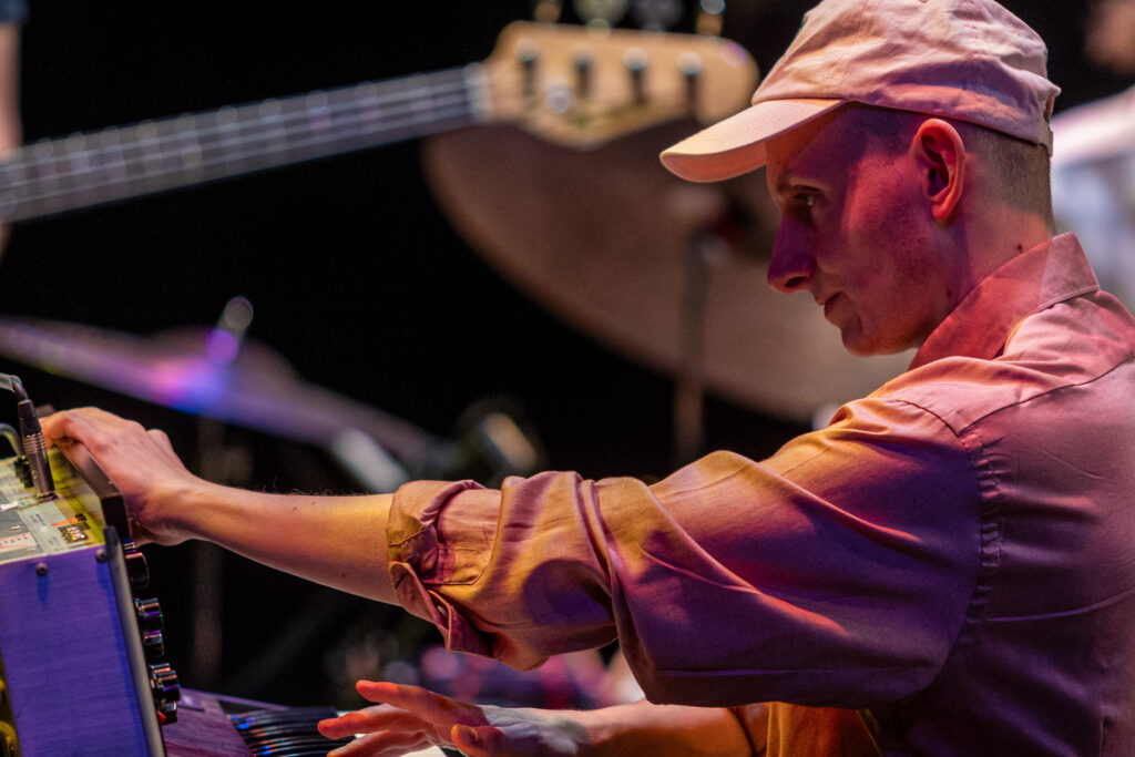 Zdjęcie. Mężczyzna w różowawej czapce z daszkiem gra na keyboardzie. Lewą ręką sięga w kierunku pokręteł na mikserze