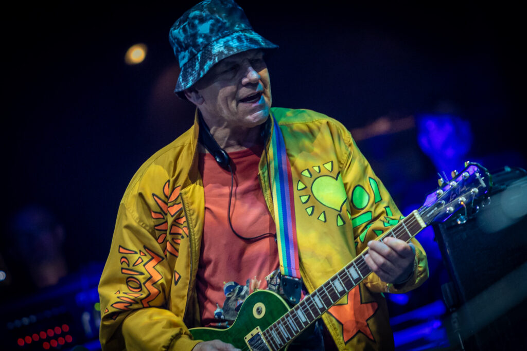 Zdjęcie. Mężczyzna w żóltej, wzorzystej koszuli i i kapeluszu na głowie gra na gitarze. To Włodzimierz Kiniorski.