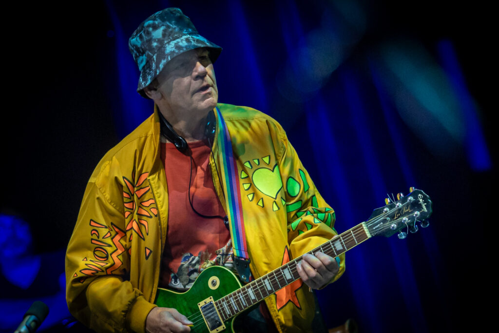 Zdjęcie. Mężczyzna w żóltej, wzorzystej koszuli i i kapeluszu na głowie gra na gitarze. To Włodzimierz Kiniorski.