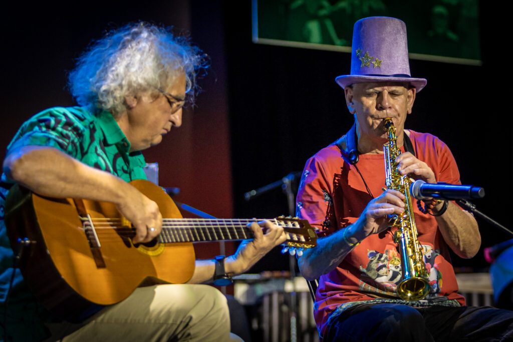 Zdjęcie. Dwaj mężczyźni siedzą na krzesłach i grają. Jeden na gitarze, drugi na klarnecie.