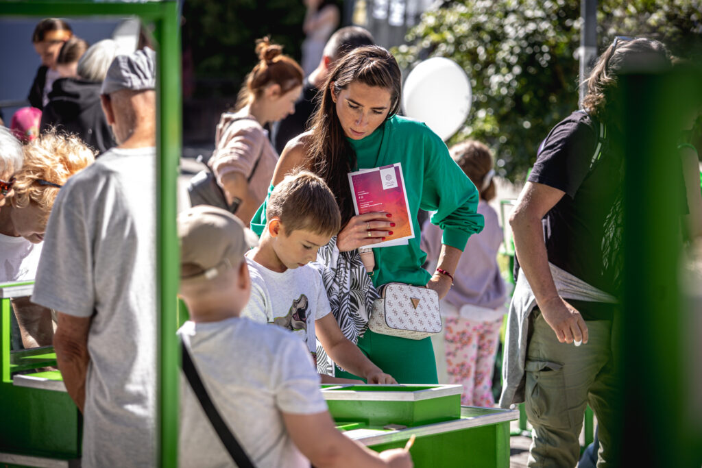 Zdjęcie. Chłopiec pochyla się nad zielonym stolikiem. Za nim stoi kobieta, która przygląda się temu co robi dziecko.