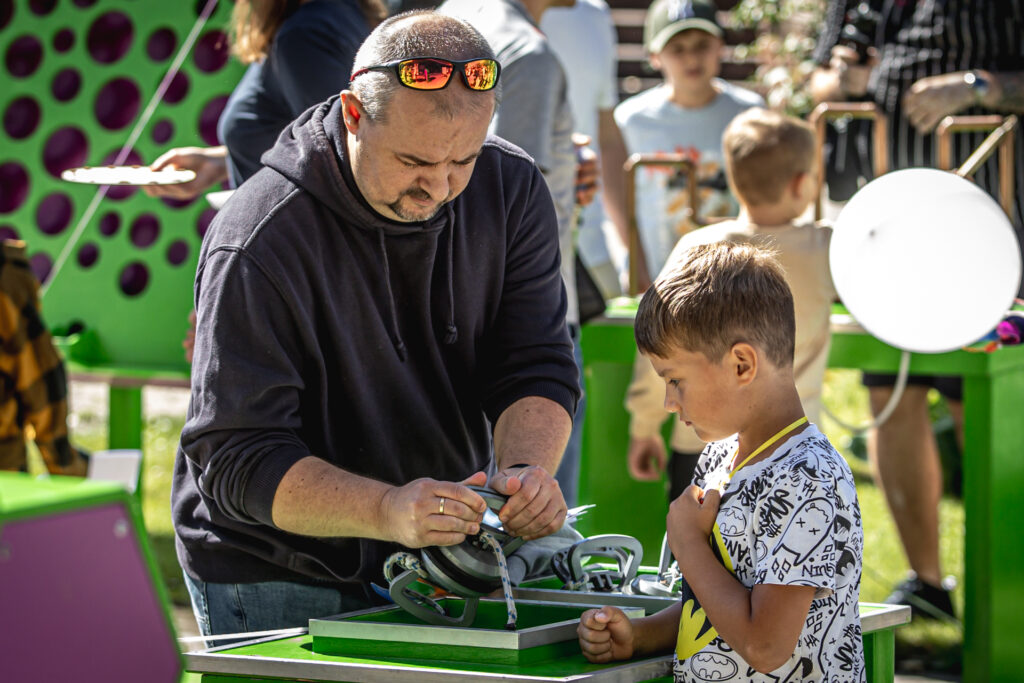 Zdjęcie. Mężczyzna i mały chłopec stoją przy zielonym stoliku. Mężczyzna ściska w rękach metalowy, duży przedmiot. Chłopiec przygląda się mu.