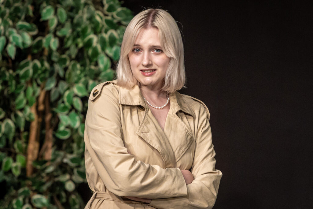 Zdjęcie. Blondynka z założonymi rękami patrzy przed siebie. Na sobie ma piaskowy płaszcz typu prochowiec.