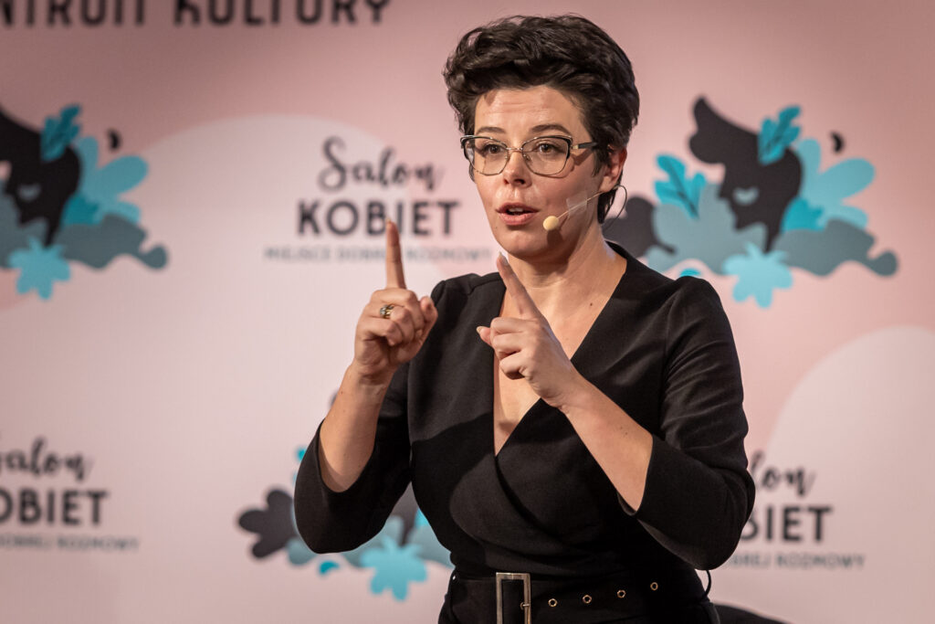 Zdjęcie. Kobieta ubrana na czarno wystawia przed siebie ręce. Miga. To Anna Kwiatkowska, tłumaczka języka migowego.