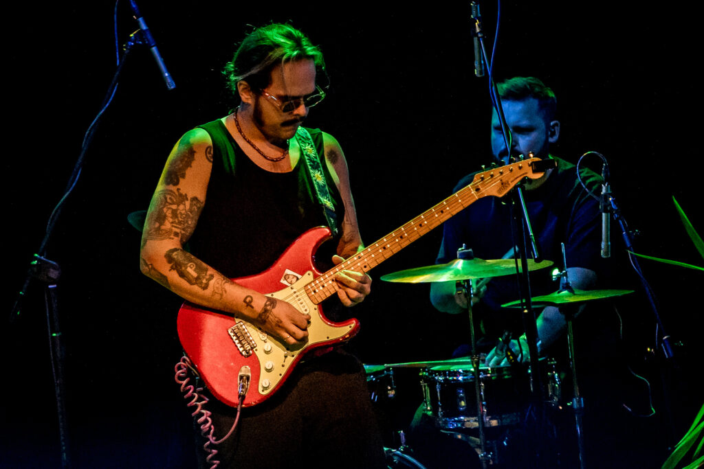 Zdjęcie. Mężczyzna gra na gitarze. W tle perkusista.