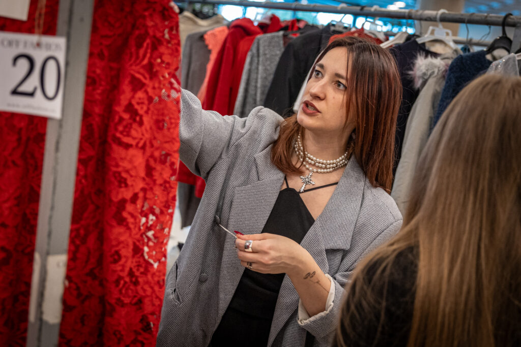 Zdjęcie. Brunetka w marynarce w drobną kratę sięgo do ubrania wiszącego na wieszaku. To czerwona koronkowa sukienka.