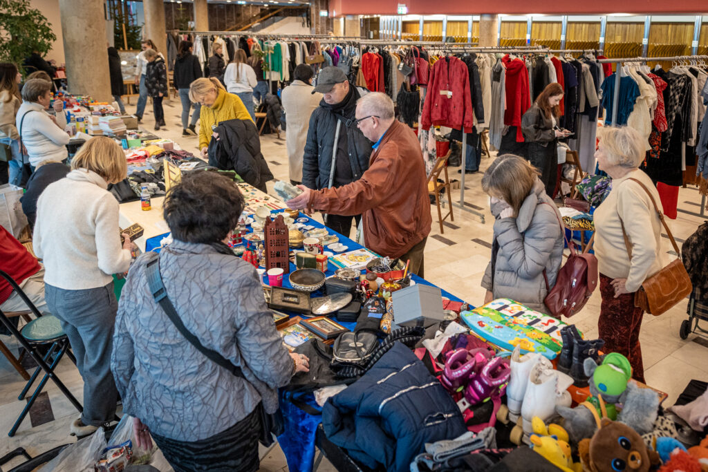 Zdjęcie. Foyer Dużej sceny. Na pierwszym planie stoły wypełnione drobnymi przedmiotami. Na drugim poustawiane w rzędy wieszaki wypełnione ubraniami.