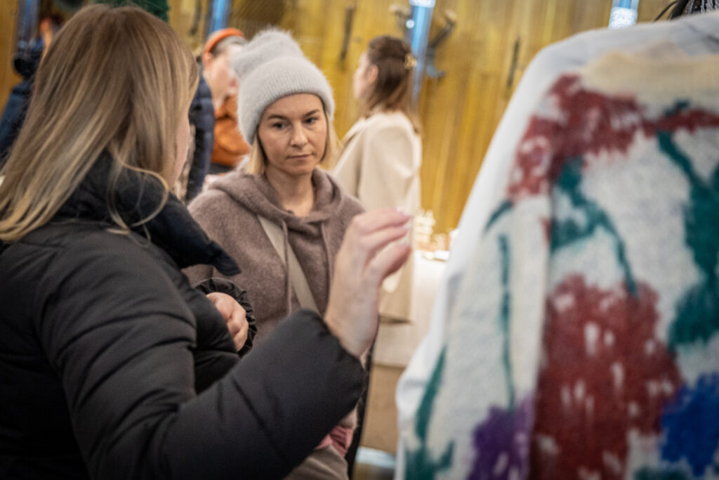Zdjęcie. Kobieta w szarej czapce przygląda się blondynce w czarnej kurtce. Ta sięga ręką w kierunku ubrania wiszącego na wieszaku.