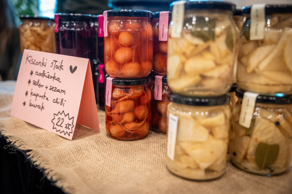 Zdjęcie. Słoiki stoące jeden na drugim przed nimi kartka kiszonki słoik: rzodkiewka, seler+sezam, kapusta chilli, burak.