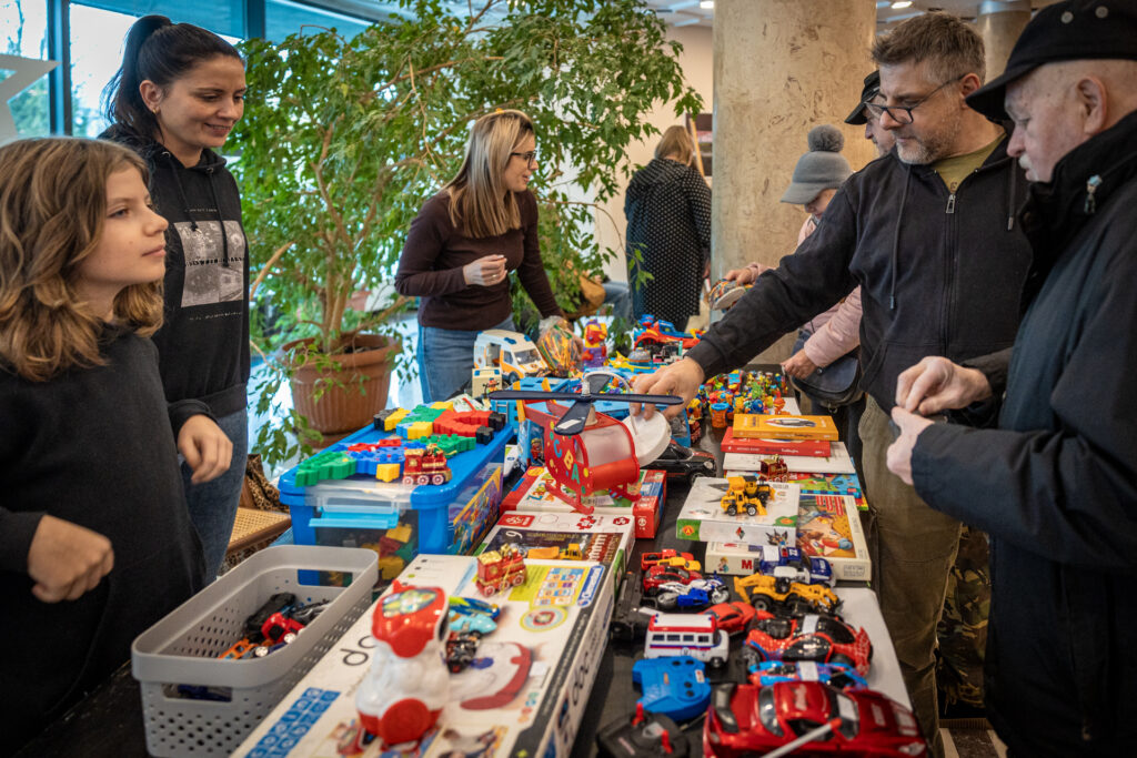 Zdjęcie. Stół zastawiony kolorowymi plastikowymi zabawkami. Mężczyźni oglądają zabawki.