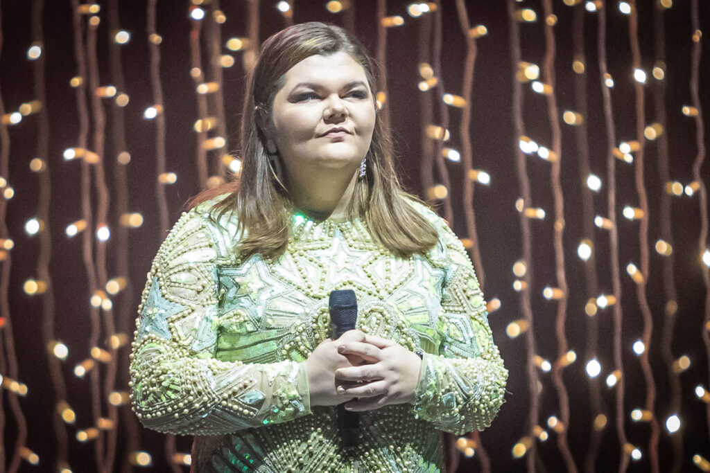 Zdjęcie. Puszysta szatynka w zielonej sukni z koralikami stoi na scenie. Trzyma mikrofon.