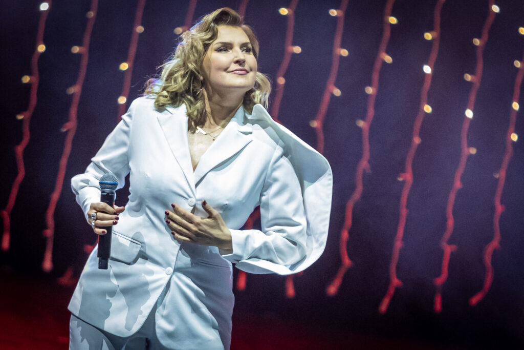Zdjęcie. Blondynka z mikrofonem na scenie. Na sobie ma biały kostium.