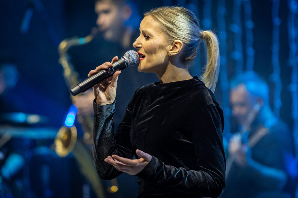 Zdjęcie. Blondynka w czarnej sukni śpiewa do mikrofonu.