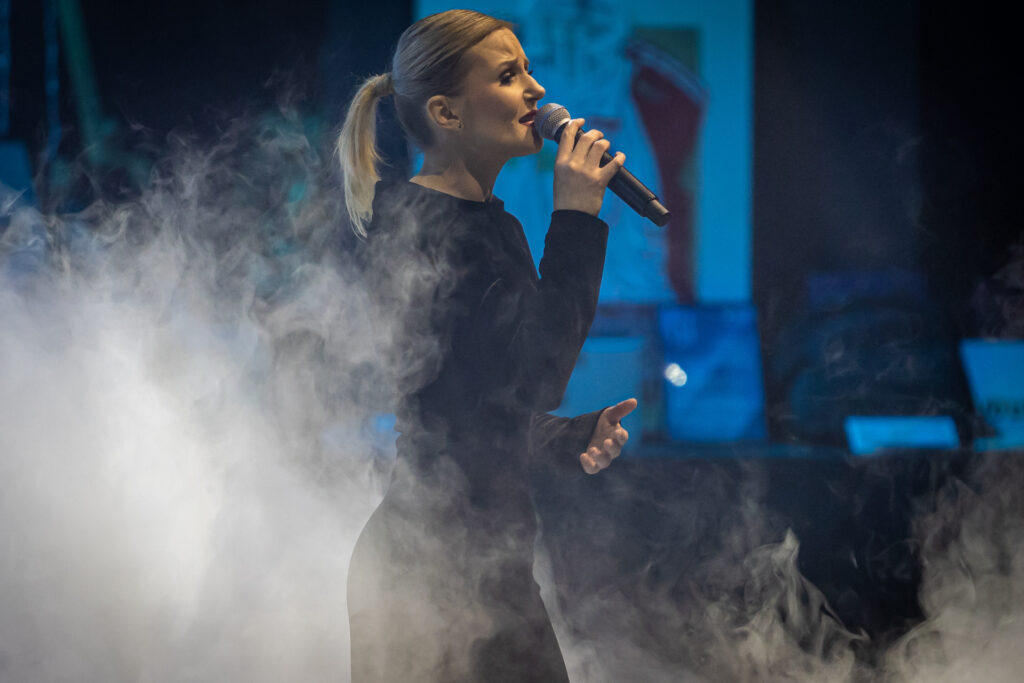 Zdjęcie. Blondynka z gładko zaczesanym kucykiem śpiewa do mikrofonu. To Kasia Lisowska.