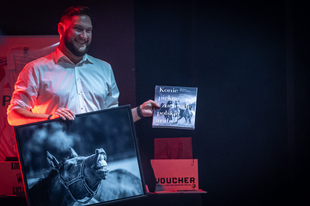 Zdjęcie. Uśmiechnięty mężczyzna trzyma w dłoni album "Konie piękne i zuchwałe. Polskie araby" oraz duże oprawione zdjęcie konia.