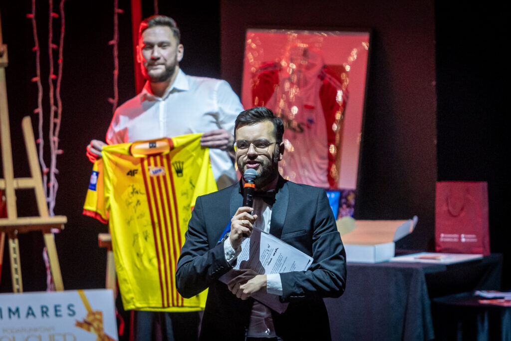 Zdjęcie. Andrzej Plata mówi do mikrofonu. W tle mężczyzna trzyma koszulkę korony Lielce a autografami.