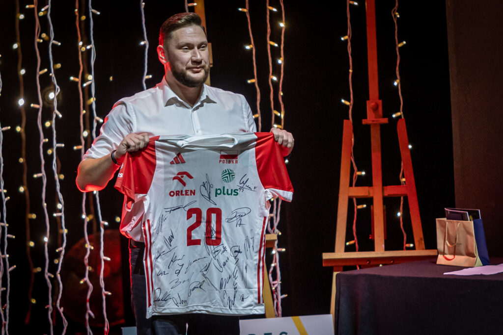 Zdjęcie. Mężczyzna trzyma podkoszulkę sportową z duża liczbą autografów. Podkoszulkajest w koloroach biało-czerwonych z flagą Polski.