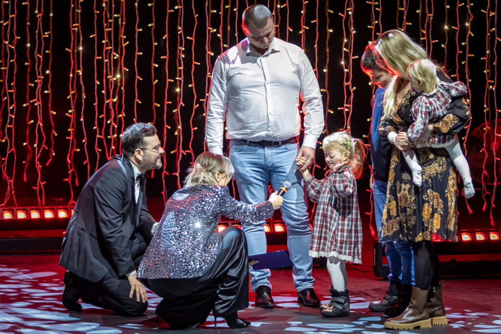 Zdjęcie. Na scenie dziewczynka w kraciastej sukience z dużym guzem w lewej gałce ocznej. Prowadząca kuca przed dziewczynką i podstawia jej mikrofon. Obok dziewczynki stoi jej rodzina: mama, tata i dwójka rodzeństwa. To Nadia Rusek i jej rodzina.