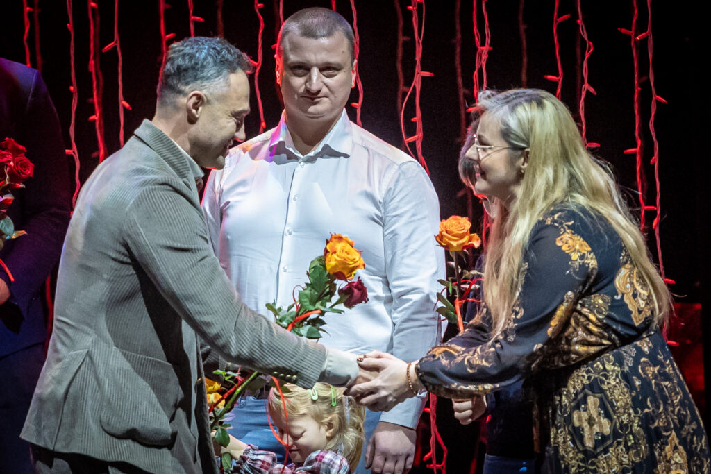 Zdjęcie. Maciej Burstein ściska dłoń mamy Nadii. Obok stoi uśmiechnięta rodzina dziewczynki.