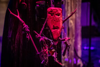 Zdjęcie. Oświetlony czerwonym reflektorem totem: zbudowany z galężi, fragmentów materiałów i z maską z oczami i ustami.
