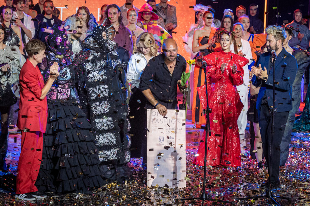 Zdjęcie. Łysy mężczyzna kłania się opierając o czek, na którym napisano: pierwsze miejsce. OFF Fashion. To Adrian Lewandowski. Po lewej stronie bije mu brawo prezydentka Kielc Agata Wojda, po prawej, wicedyrektor KCK Artur Wijata. W tle grupa projektantów i modeli. Z góry leci kolorowe, błyszczące konfetti.