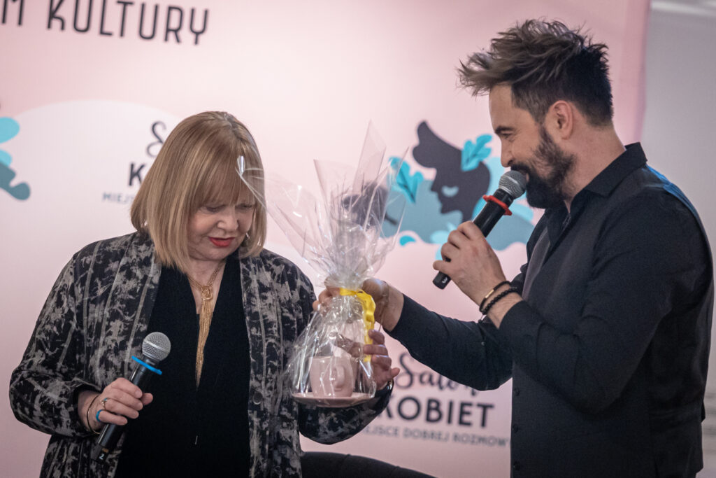Zdjęcie. Mężczyzna w ciemnej kamizelce Artur Wijata wręcza różową filiżankę Joannie Żółkowskiej.