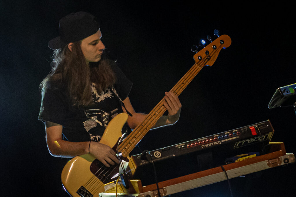 Zdjęcie. Młody mężczyzna gra na gitarze. Ma długie, proste ciemne włosy i czapkę z daszkiem założoną tyłem do przodu.