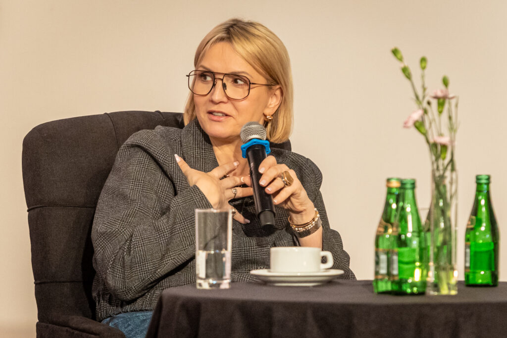 Zdjęcie. Blondynka siedzi na krześle przy stoliku. Prawą rękę przykłada do dekoltu. W lewej trzyma mikrofon i patrzy w bok. To Marta Kuligowska, dziennikarka TVN24.