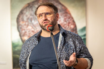 Zdjęcie. Mężczyzna mówi do mikrofonu. Ma okulary, wąsy i brodę. Ubrany w granatową podkoszulkę i rozpiętą koszulę w granatowe kwiaty. To Bartłomiej Radosz.