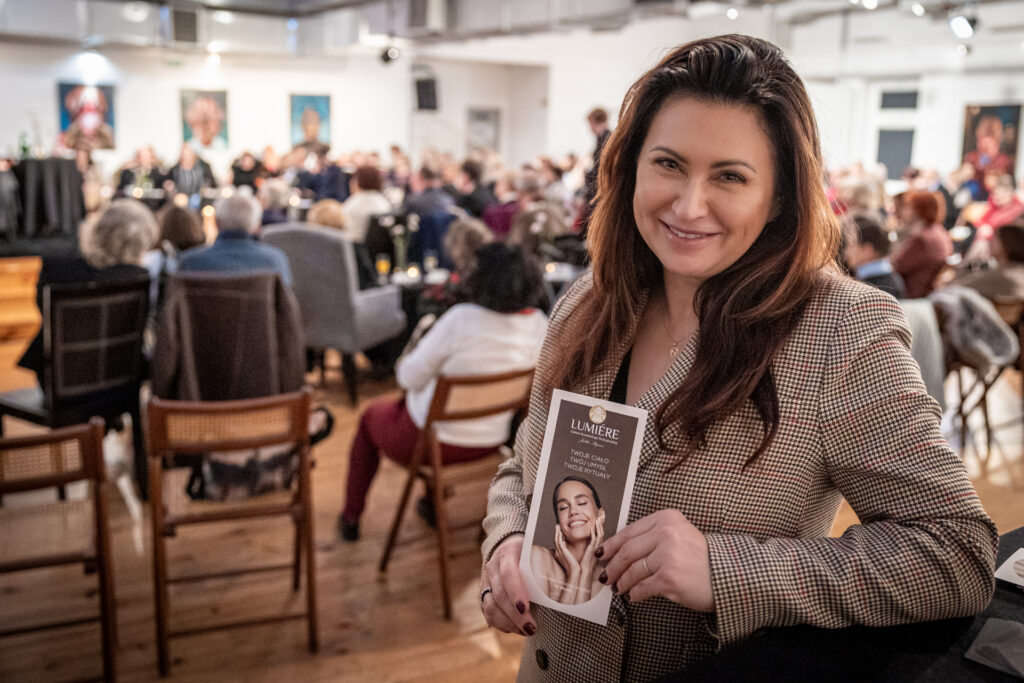 Zdjęcie. Uśmiechnięta kobieta pozuje do zdjęcia. W dłoniach trzyma ulotkę reklamową salonu Lumiere. To Julita Stępień, szefowa gabinetu kosmetycznego.
