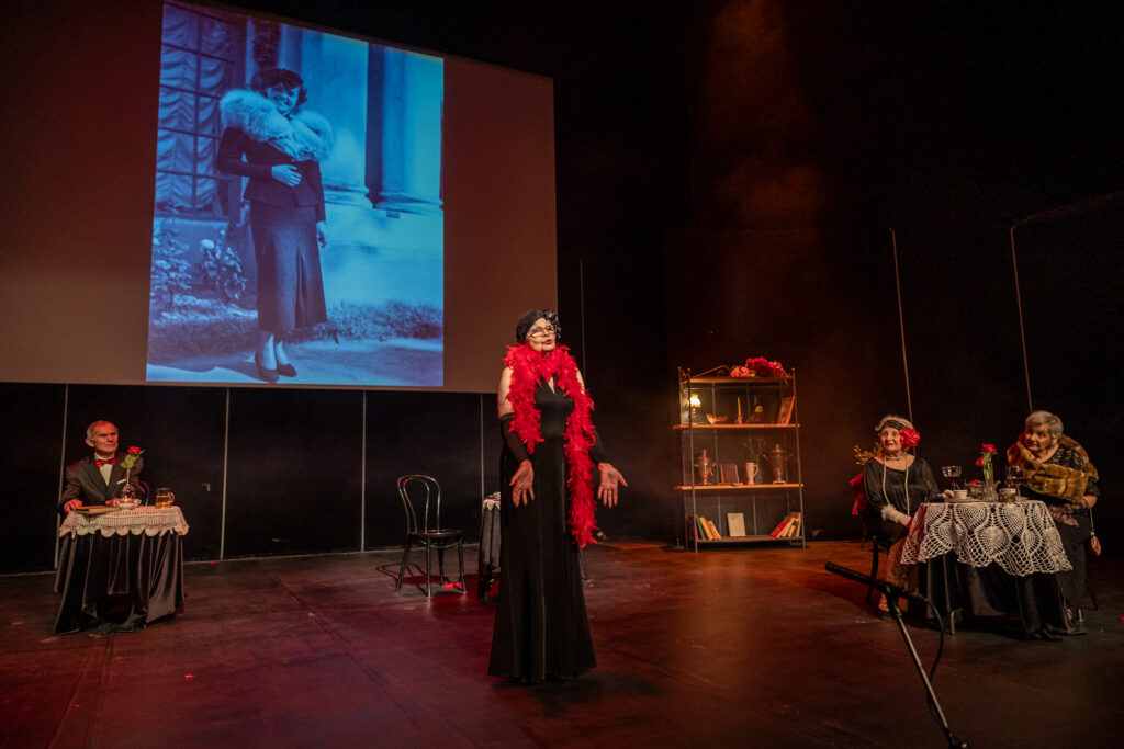 Zdjęcie. Mała Scena KCK podczas spektaklu Teatru bez Suflera. N środku stoi kobieta z czerwonym boa. Dookoła przy stolikach siedzą inni aktorzy