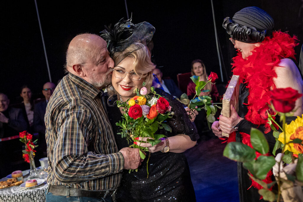 Zdjęcie. Starszy łysy mężczyzna w koszuli w kratkę przytula i całuje w policzek uśmiechniętą aktorkę Teatru bez Suflera.