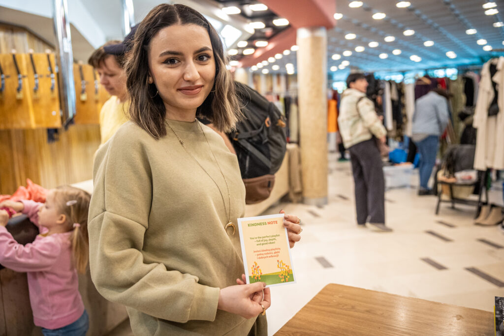 Zdjęcie. Młoda brunetka prezentuje karteczkę-pocztówkę wyprzedaży z optymistycznym cytatem.