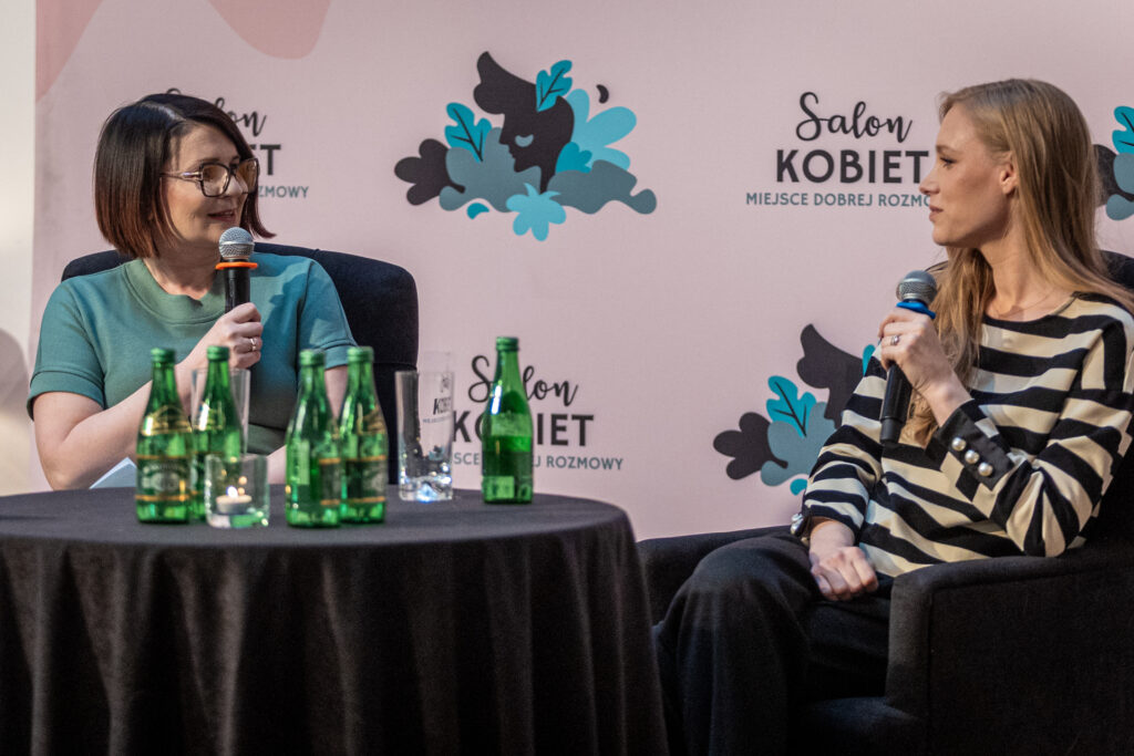 Zdjęcie. Dwie kobiety siedzą przy okrągłym stoliku z mikrofonami. Jedna to brunetka w okularach - prowadząca spotkanie Monika Sierpień, druga to blondynka o długich prostych włosach - to gościni spotkania Magda Łucyan.