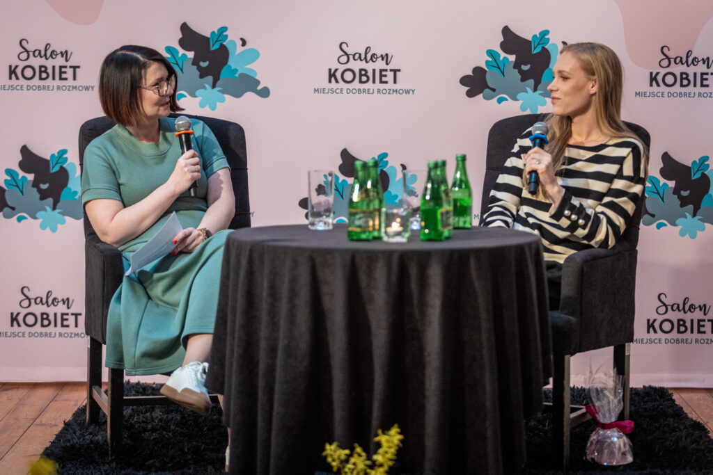 Zdjęcie. Dwie kobiety siedzą przy okrągłym stoliku. W dłoniach trzymają mikrofony. Za nimi różowa ścianka z napisem Salon Kobiet.