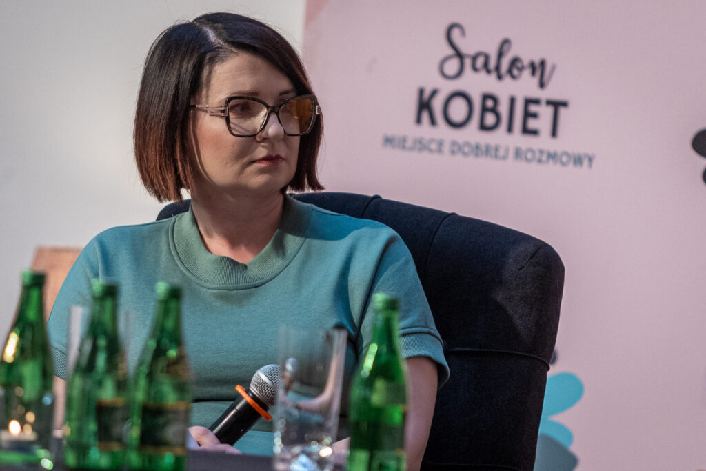 Zdjęcie. Brunetka o prostych włosach za ucho patrzy w bok. To prowadząca spotkanie Monika Sierpień.