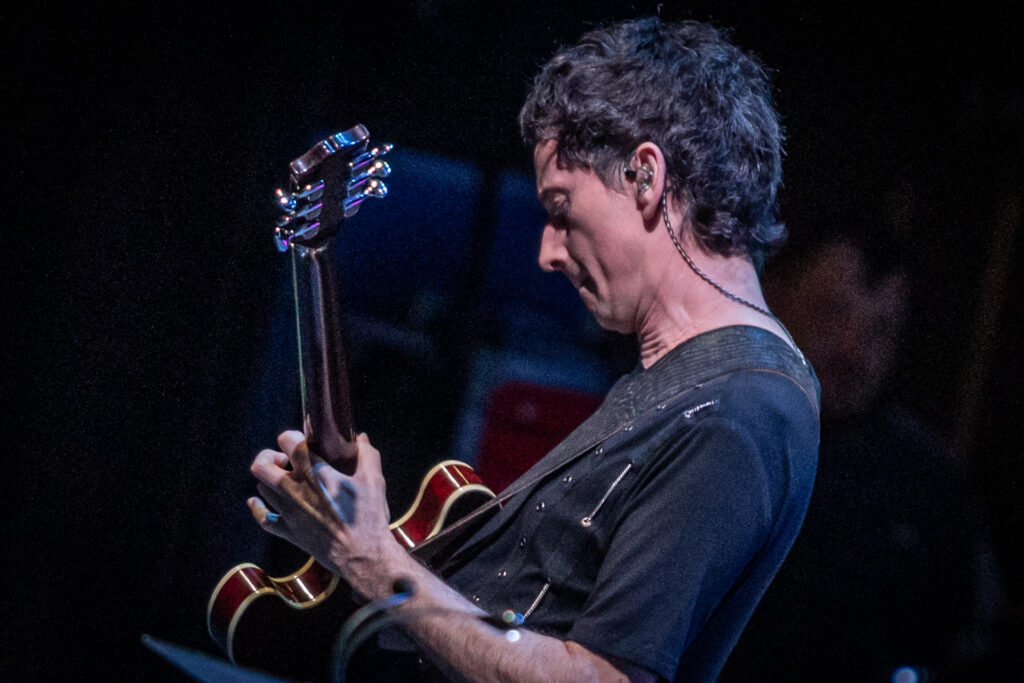 Zdjęcie. Mężczyzna gra na gitarze. W uchu ma słuchawkę z odchodzącym od niej kablem.