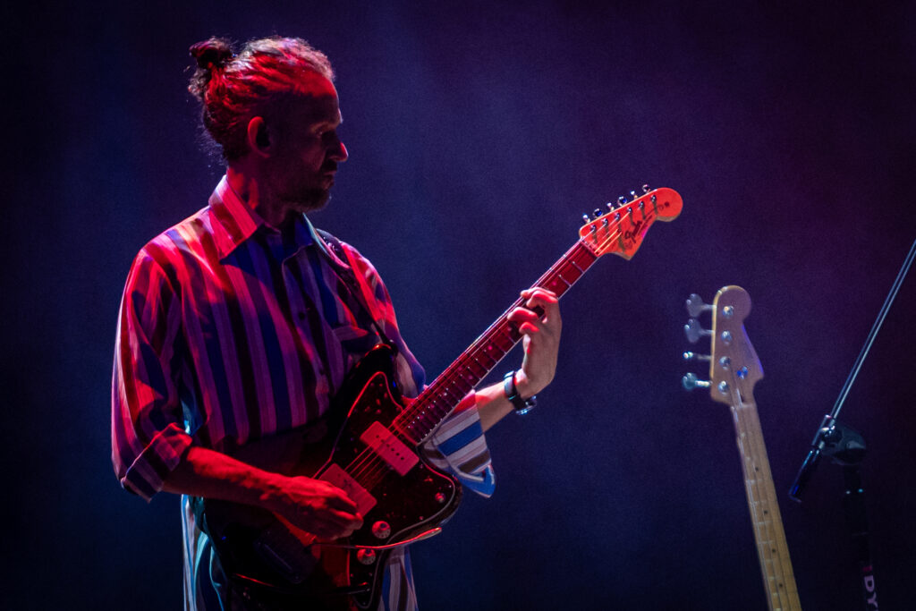 Zdjęcie. Mężczyzna z dłuższymi włosami spiętymi w kucyk gra na gitarze.