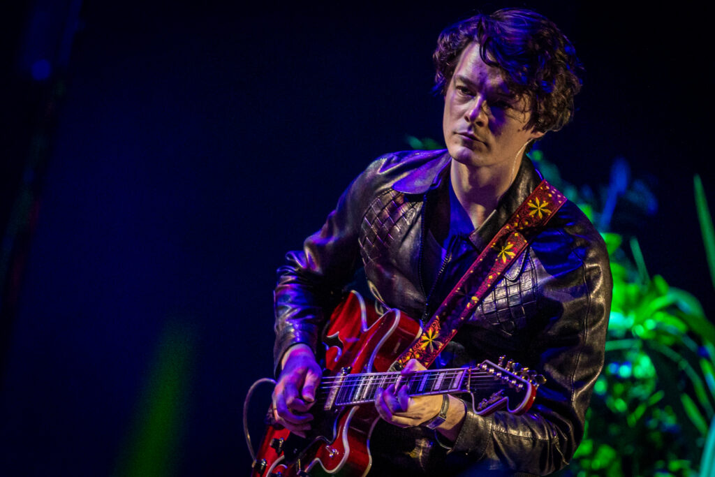 Zdjęcie. Tomasz Ziętek gra na gitarze. To mężczyzna o ciemnych dłuższych włosach z grzywką. Ma skórzaną dopasowaną kurtkę z kołnierzem.