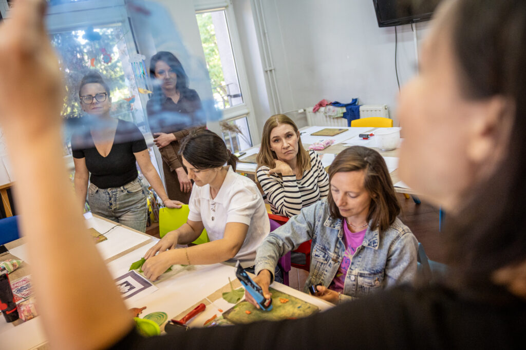 Zdjęcie. Kobiety w pracowni plastycznej patrzą na podnoszącą do góry szablon prowadzącą warsztaty.