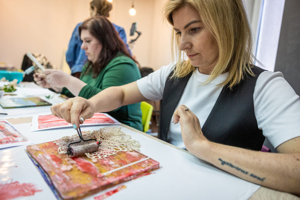 Zdjęcie. Blondynka jeździ białym wałkiem malarskim po serwetce rozłożonej na płytce.