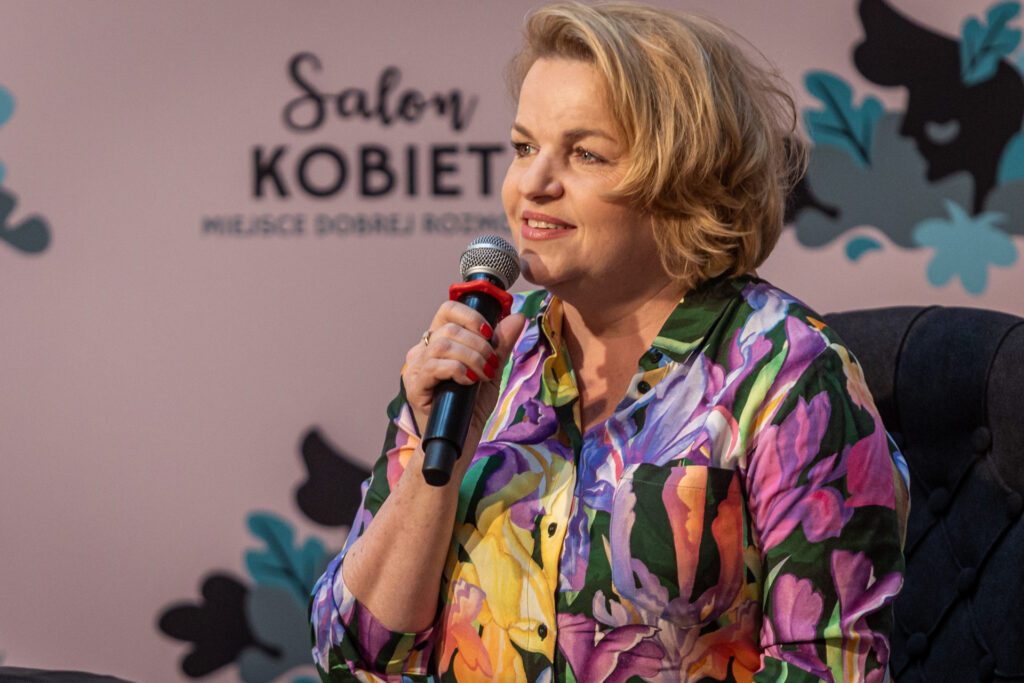 Zdjęcie. Kobieta w kwiecistej koszuli mówi do mikrofonu. To Katarzyna Bosacka.