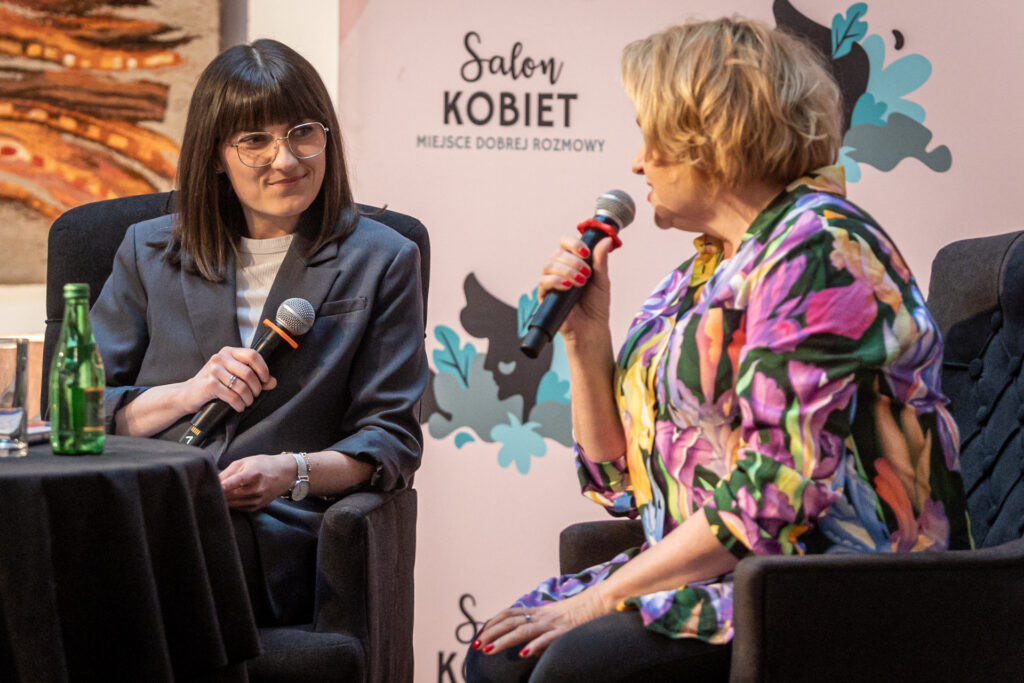 Zdjęcie. Dwie kobiety z mikrofonami siedzą przy stoliku. Anita Michalczyk - prowadząca - uśmiecha się. Do mikrofonu mówi Katarzyna Bosacka.