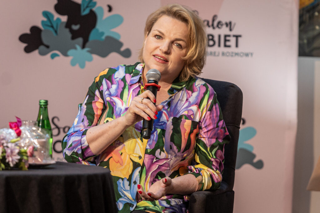 Zdjęcie. Kobieta w kwiecistej koszuli mówi do mikrofonu. To Katarzyna Bosacka.