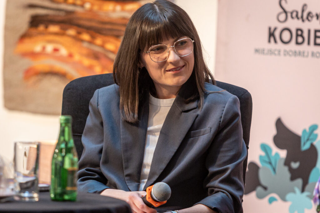 Zdjęcie. Kobieta siedzi na fotelu. W dłoni trzyma mikrofon. To brunetka o prostych włosach poniżej ramion i grzywce. Kobieta uśmiecha się. To Anita Michalczyk.