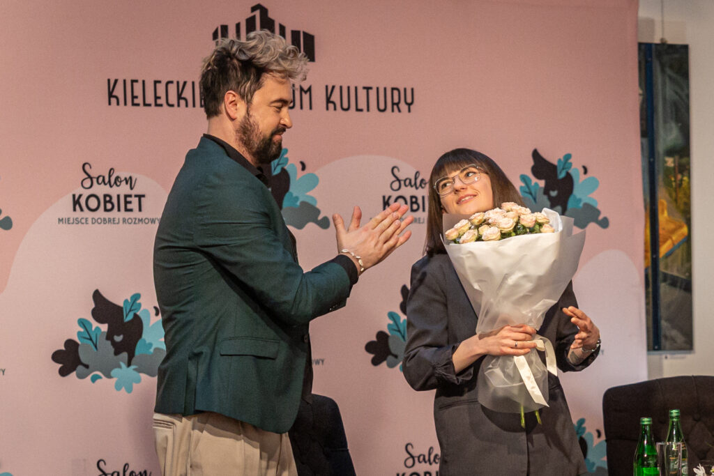 Zdjęcie. Mężczyzna o jasnej grzywce z ciemną brodą klaszcze patrząc w kierunku niewysokiej kobiety w okularach. Ta trzyma w dłoni bukiet białych róż. To p.o. dyrektora KCK Artur Wijata i prowadząca Salon Kobiet Anita Michalczyk.